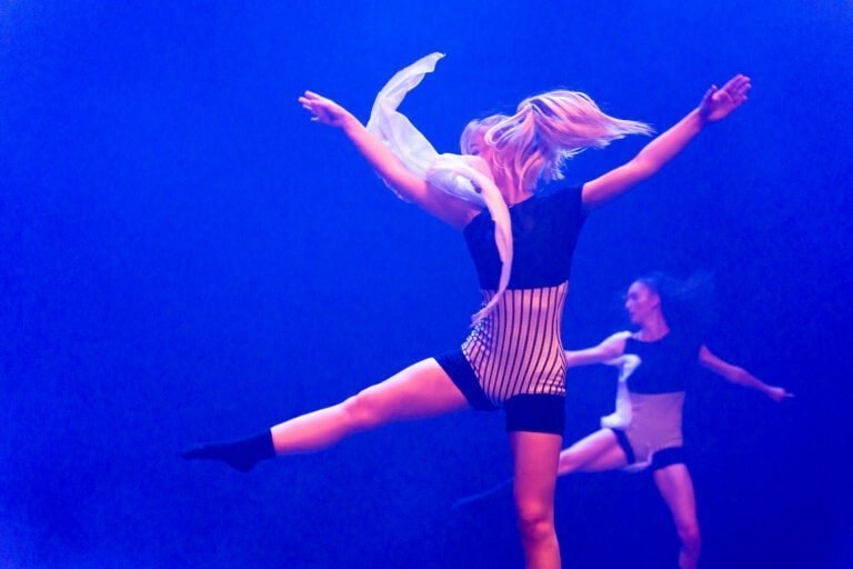 Deux danseuses exécutent une routine dynamique sur scène, éclairée par un éclairage bleu. Une danseuse au premier plan saute, les bras tendus et les cheveux ondulants, portant des chaussettes noires et une tenue rayée. La deuxième danseuse est légèrement floue à l'arrière-plan, reflétant un mouvement similaire au Spectacle de danse à Floirac.