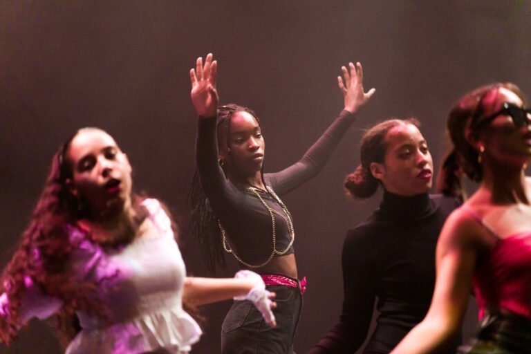 Un groupe de quatre femmes se produit sur scène, dansant de manière expressive, les bras levés et vêtues de diverses tenues de couleur sombre. L'arrière-plan est faiblement éclairé par des teintes roses et violettes, créant une atmosphère dramatique. Dans ce Spectacle de danse à Floirac, l'attention est centrée sur la femme au centre, qui lève les mains avec assurance.