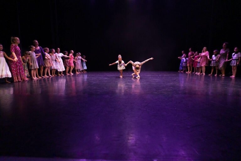 Deux enfants exécutent une chorégraphie sur une scène faiblement éclairée du Spectacle de danse à Floirac, chacun prenant la pose avec un bras tendu. Ils sont encadrés par un demi-cercle d'autres enfants qui les regardent depuis le bord de la scène, vêtus de divers costumes colorés.