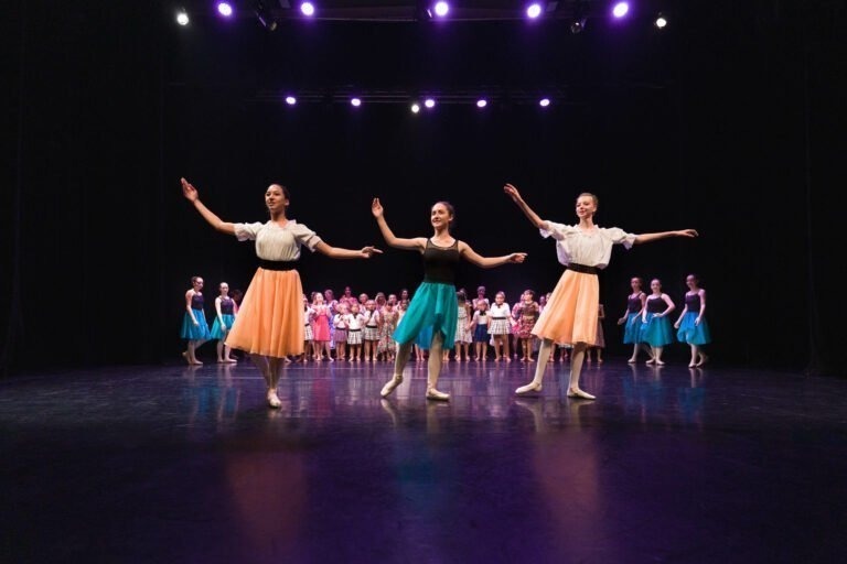 Trois danseurs de ballet se produisent sur scène sous des lumières violettes, les bras gracieusement étendus. Vêtus de hauts blancs et de jupes colorées orange et bleu canard sur un fond sombre, ils captivent le public. Derrière eux, des danseurs aux costumes divers assistent au spectacle de danse à Floirac.