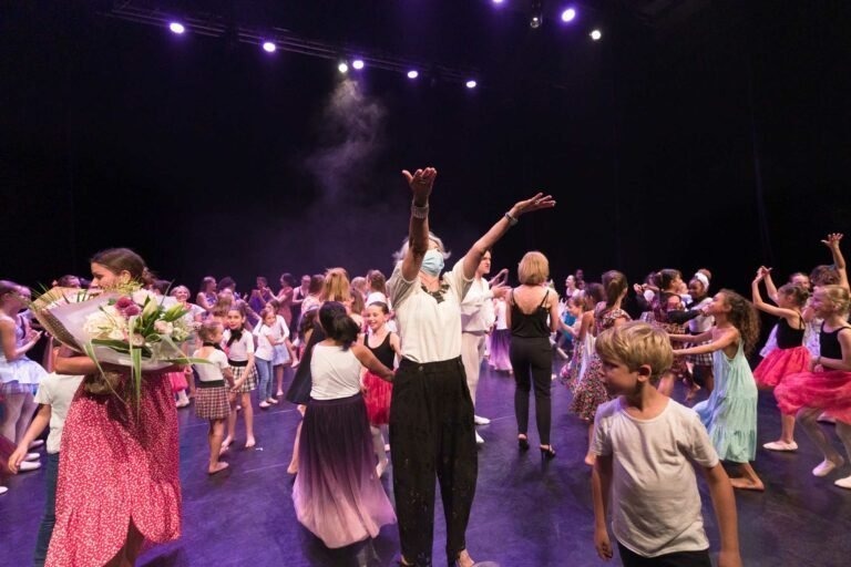 Un groupe d'enfants et quelques adultes sont réunis sur une scène sombre sous des lumières violettes au Spectacle de danse à Floirac. Certains enfants portent des costumes, notamment des tutus, tandis que d'autres sont en tenue décontractée. Une personne au centre tend les bras vers le haut, tandis qu'une autre tient un bouquet de fleurs.