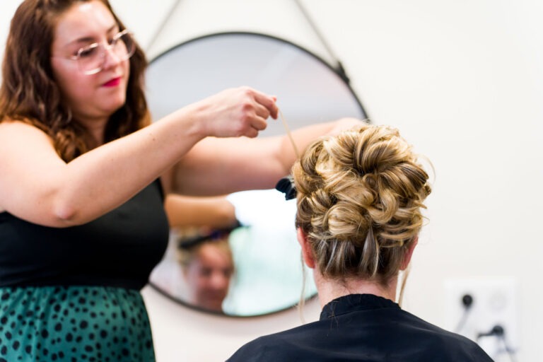 Un coiffeur aux cheveux ondulés, portant des lunettes et un haut noir avec une jupe à pois verts, coiffe une femme blonde devant un miroir rond. En préparation pour son mariage, la femme assise sur la chaise dévoile des boucles complexes tandis que le miroir reflète les deux femmes.