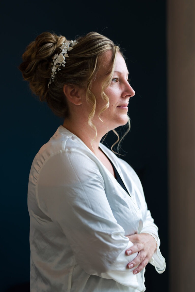 Une femme aux cheveux châtain clair et bouclés, coiffés en chignon élégant orné d'accessoires floraux blancs, se tient debout, les bras croisés, regardant par la fenêtre. Elle porte un chemisier clair et semble plongée dans ses pensées, comme si elle réfléchissait à son mariage à venir, éclairée par la douce lumière naturelle de la fenêtre.