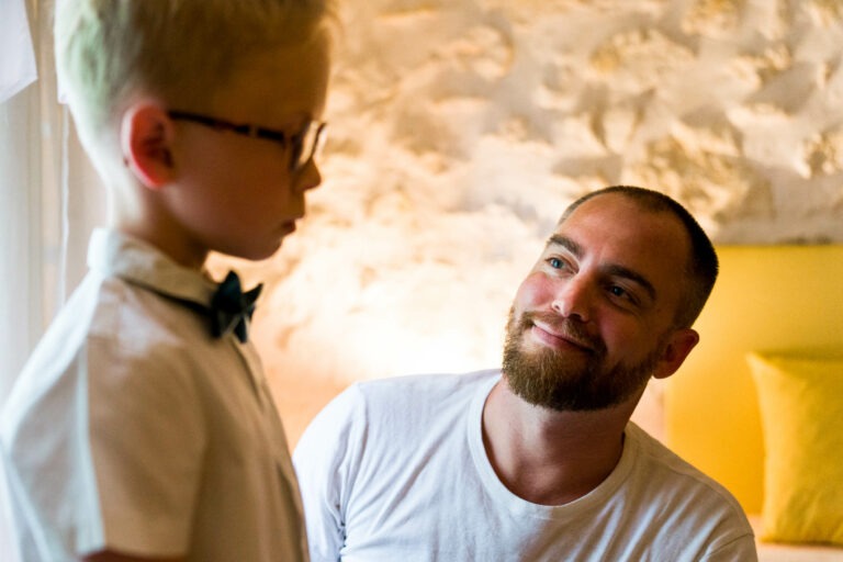 Un homme barbu au crâne rasé, vêtu d'un t-shirt blanc, sourit chaleureusement à un enfant portant des lunettes, une chemise blanche et un nœud papillon. Ils sont à l'intérieur, dans une pièce bien éclairée aux murs rustiques en pierre blanche et aux coussins jaune vif. La scène évoque le charme des mariages bordeaux, mêlant élégance et chaleur.