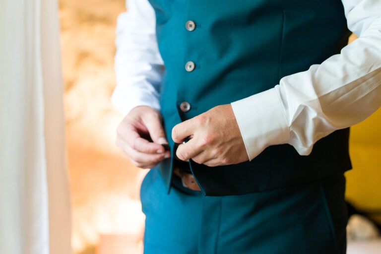 Une personne vêtue d'une chemise blanche boutonne le devant d'un gilet bleu canard, rappelant une tenue raffinée pour un mariage. L'accent est mis sur ses mains et son torse. L'arrière-plan est flou, mettant en valeur une ambiance chaleureuse et doucement éclairée avec des touches de tons beiges et jaunes.