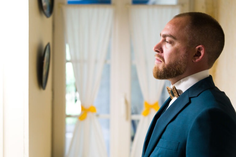 Un homme barbu, vêtu d'un costume bleu et d'un nœud papillon, se tient de profil à l'intérieur, tourné vers la droite. Il semble être plongé dans ses pensées ou dans sa réflexion, peut-être à propos d'un mariage à venir. Derrière lui, des rideaux blancs avec des rubans jaunes noués en nœuds laissent entrer la lumière naturelle à travers de grandes fenêtres. Les murs sont décorés d'assiettes suspendues.