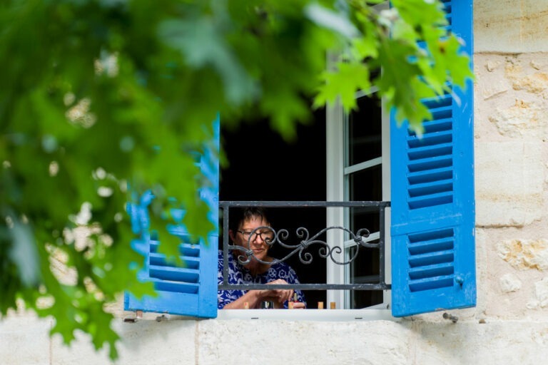 Une personne portant des lunettes regarde avec satisfaction par une fenêtre encadrée de volets bleu vif. L'individu semble tenir un verre de Bordeaux tout en portant une chemise à motifs floraux. Des feuilles vertes luxuriantes obscurcissent partiellement la vue, ajoutant une touche naturelle à la scène. La fenêtre est encastrée dans un mur en pierre claire.