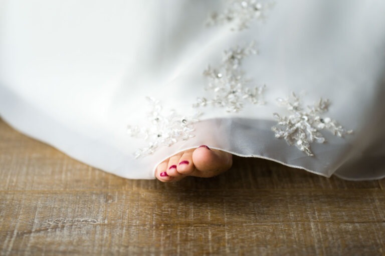 Une image en gros plan montre un pied aux ongles peints en rouge qui dépasse d'une robe de mariée blanche ornée de dentelle. La robe est ornée de délicats détails en dentelle florale et le pied, rappelant l'élégance des mariages bordelais, repose sur un plancher en bois.