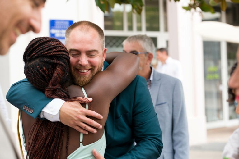 Un homme barbu portant une veste bleu canard et une chemise blanche serre chaleureusement dans ses bras une femme aux cheveux tressés et portant une robe sans manches bleu clair. Ils sont dehors pour ce qui semble être un mariage joyeux, avec des personnes floues en arrière-plan, dont un homme en costume gris. Tout le monde semble sourire et profiter du moment.