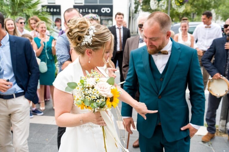 Un couple de mariés est dehors, entouré d'invités qui applaudissent. La mariée, en robe blanche, tient un bouquet de tournesols et de roses blanches, pointant du doigt le costume vert du marié. Le marié porte un nœud papillon et une barbe, regardant vers le bas dans la direction qu'elle pointe du doigt. L'ambiance est joyeuse et festive, comme lors d'un mariage élégant à Bordeaux.