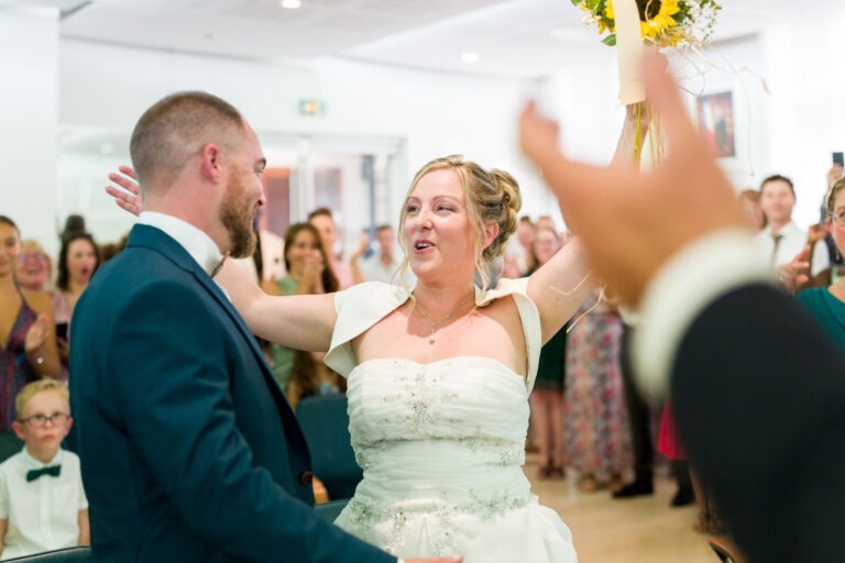 Un couple de jeunes mariés se tient dans une salle lumineuse remplie d'invités. La mariée, vêtue d'une robe blanche, lève les bras joyeusement tout en tenant des tournesols. Le marié, en costume sombre, lui sourit. Les invités derrière eux semblent applaudir et célébrer. Une main floue est visible au premier plan de leur magnifique mariage bordeaux.