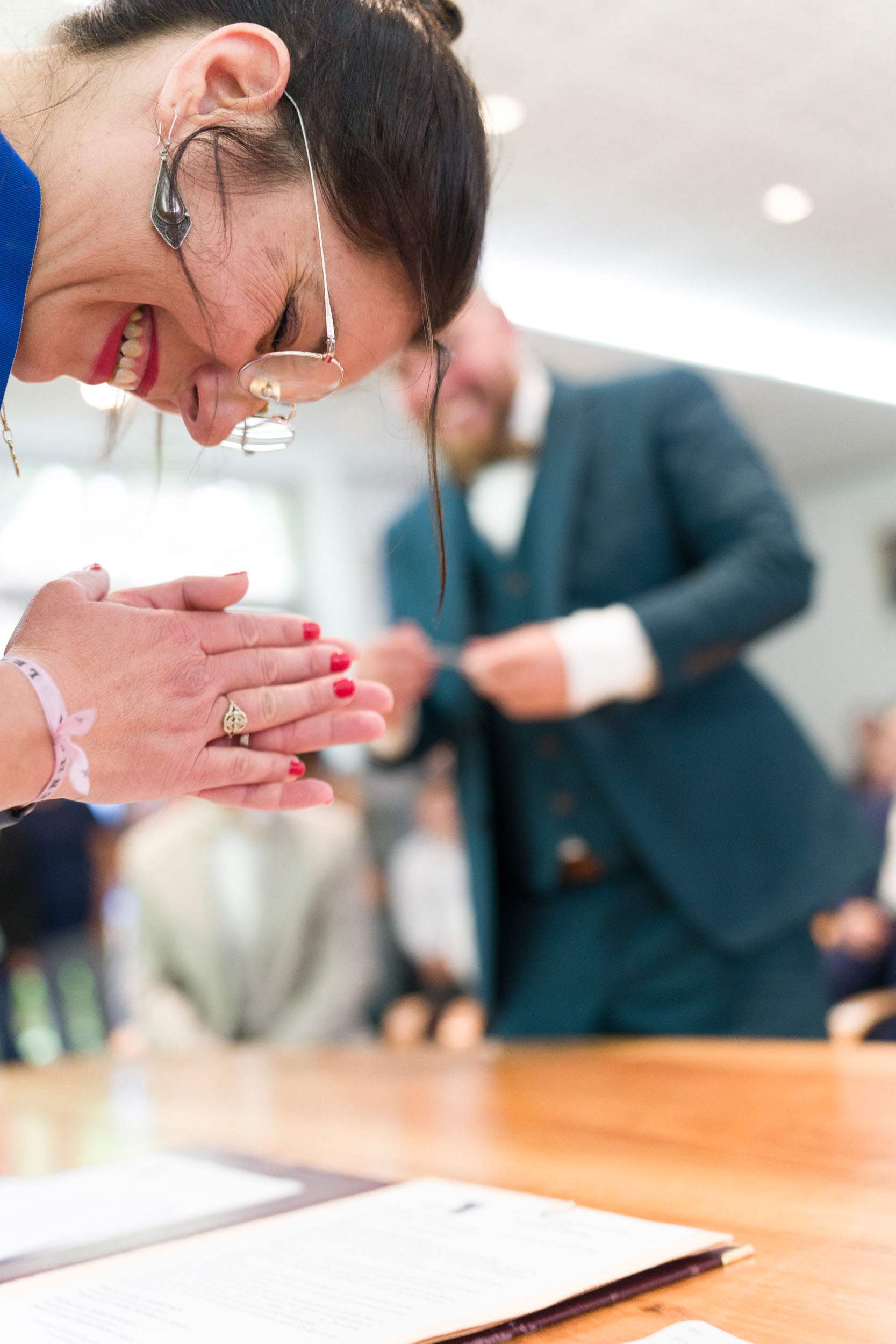 Une femme portant un haut bleu sourit et joint les mains en signe de gratitude, probablement lors d'une célébration de mariage. Elle porte des lunettes et a les ongles peints en rouge. Un homme en costume est visible à l'arrière-plan, légèrement flou. Ils se trouvent dans un décor intérieur très éclairé avec une table en bois clair au premier plan.