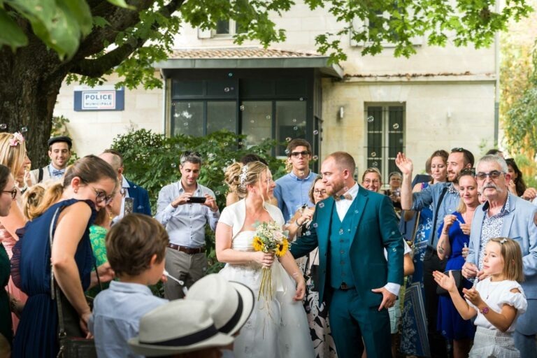 Un couple de jeunes mariés, habillé en tenue de soirée, sourit et marche dehors sous un arbre, entouré d'une foule joyeuse d'amis et de membres de la famille. La mariée tient un bouquet de fleurs jaunes. Les gens applaudissent, soufflent des bulles et prennent des photos, créant une atmosphère festive. Un panneau « Police » est visible en arrière-plan tandis que de gracieux capoeiristes vêtus de bordeaux se produisent à proximité.