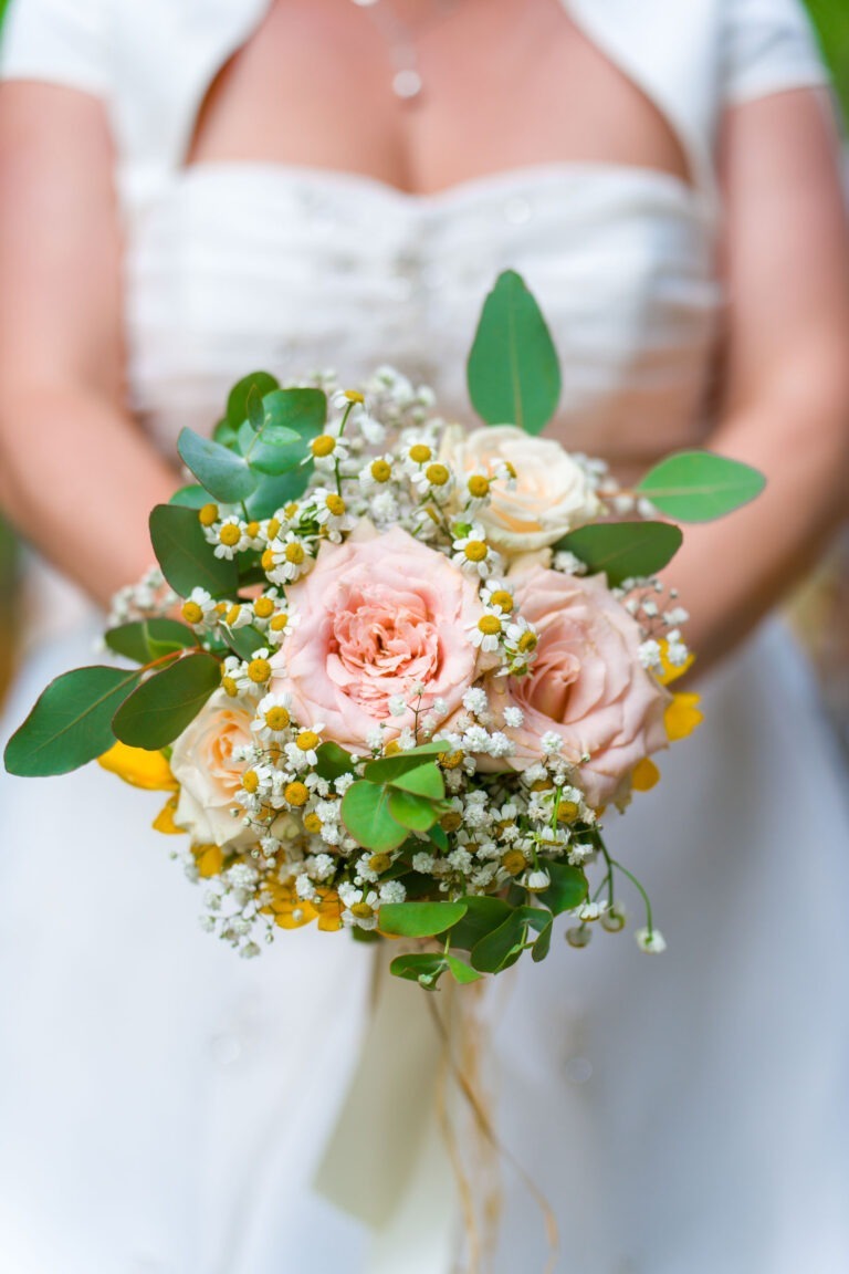 Une mariée en robe blanche tient un bouquet de roses roses et blanches, de petites fleurs jaunes, de gypsophile et de feuillage vert. L'arrière-plan est flou, attirant l'attention sur le bouquet et une partie du haut du corps de la mariée pendant ce mariage pittoresque.