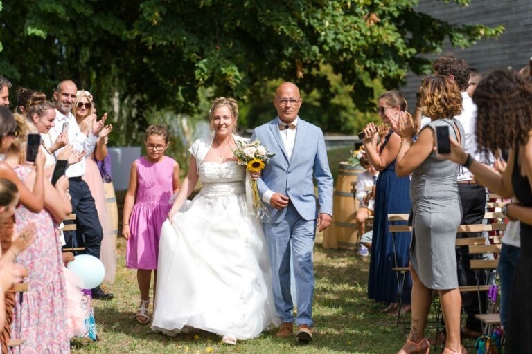 Une mariée en robe blanche et un marié en costume bleu clair marchent dans une allée extérieure en souriant. Ils sont entourés d'invités qui les applaudissent, dont une jeune fille en robe rose tenant la main de la mariée. Le cadre est verdoyant et des chaises en bois bordent l'allée lors de ce mariage sur le thème du bordeaux.