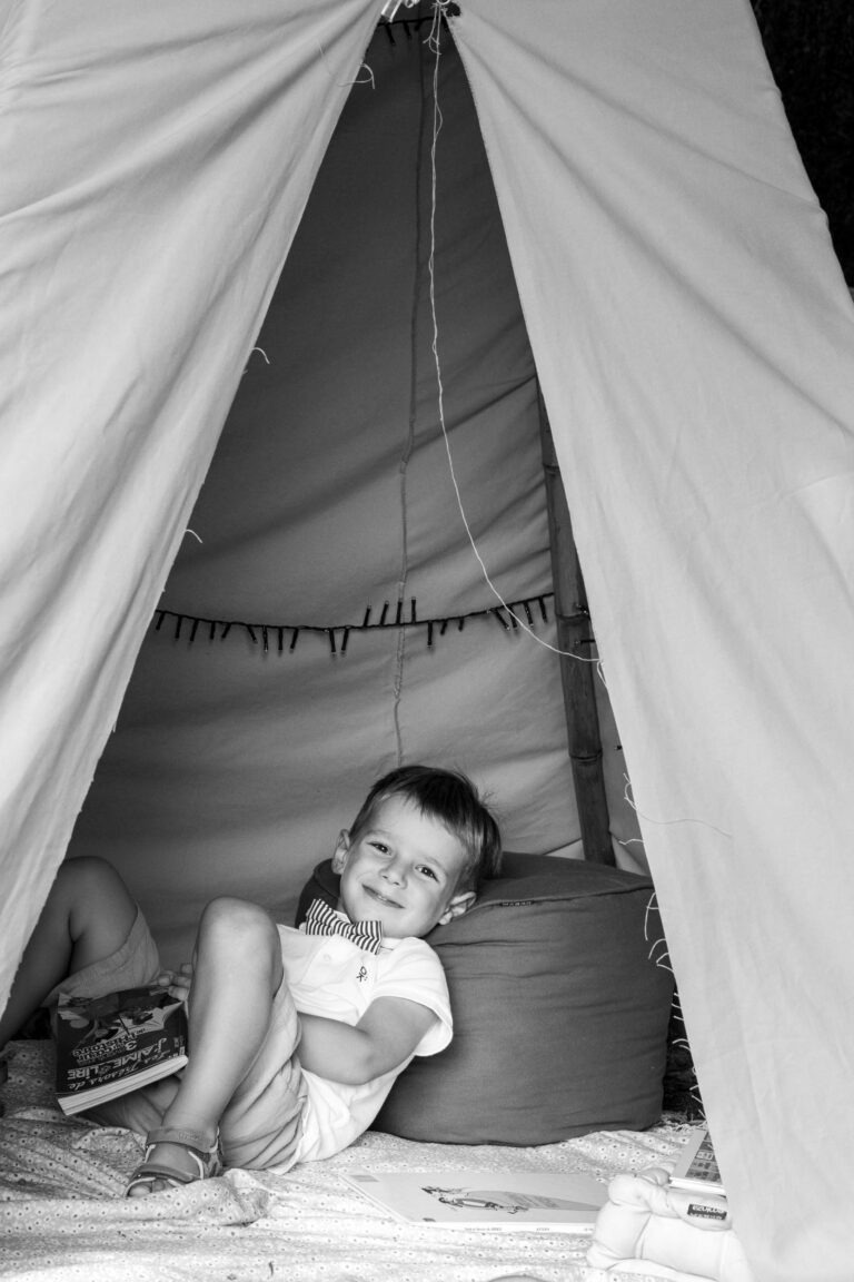 Image en noir et blanc d'un jeune enfant assis dans une petite tente tipi. L'enfant tient un livre et sourit tout en s'appuyant sur un coussin. L'ouverture de la tente révèle un intérieur douillet avec une autre personne partiellement visible, allongée avec seulement ses jambes apparentes. Divers objets, dont du matériel de capoeiriste, sont éparpillés à l'extérieur de la tente.