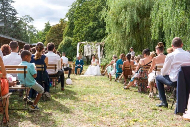 Une cérémonie de mariage en plein air avec des invités assis sur des chaises en bois disposées en deux rangées face à une tonnelle décorative. Les mariés sont assis à l'avant sous la tonnelle ornée de tissu blanc et de verdure. Le décor est un espace gazonné entouré d'arbres sous un ciel partiellement nuageux, créant une atmosphère bordelaise enchanteresse.