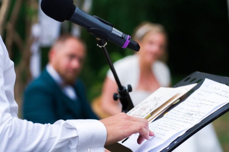 Gros plan d'une personne parlant dans un micro, avec des notes sur un support devant elle. En arrière-plan, légèrement flou, on voit un homme en costume bleu canard et une femme en robe blanche, suggérant le mariage. L'accent est mis sur la main de l'orateur qui pointe les notes.