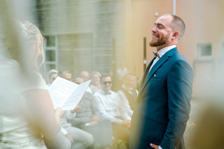 Un marié en costume bleu et nœud papillon sourit face à sa future épouse, qui lit un papier, lors d'une cérémonie de mariage en plein air. Le premier plan flou ajoute une touche de douceur et d'émotion. Des invités en vêtements de couleur claire et lunettes de soleil sont assis à l'arrière-plan devant un bâtiment moderne à Bordeaux.