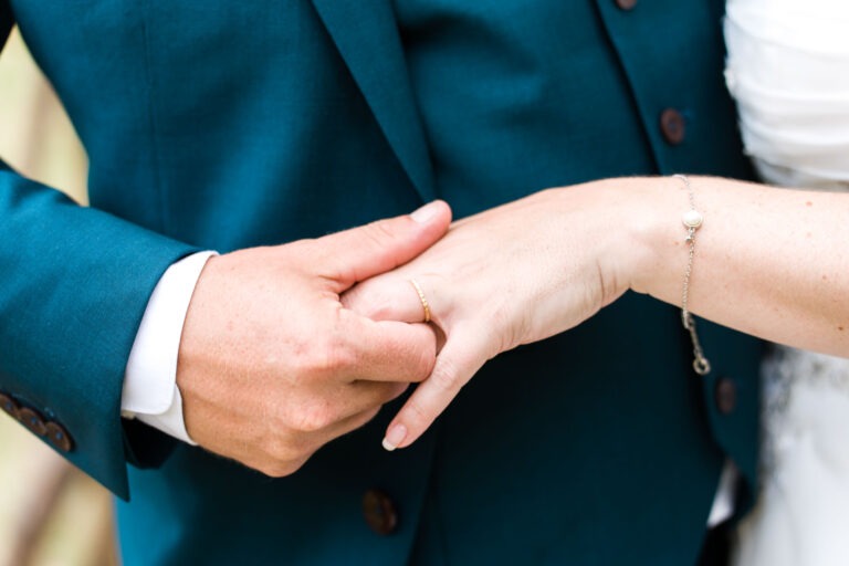 Gros plan d'un couple le jour de leur mariage, le marié en costume bleu canard tenant la main de la mariée. La robe blanche de la mariée et un bracelet délicat avec une seule perle sont partiellement visibles. La main de la mariée porte une bague en or, symbolisant leur lien conjugal alors qu'ils célèbrent leur mariage à Bordeaux.