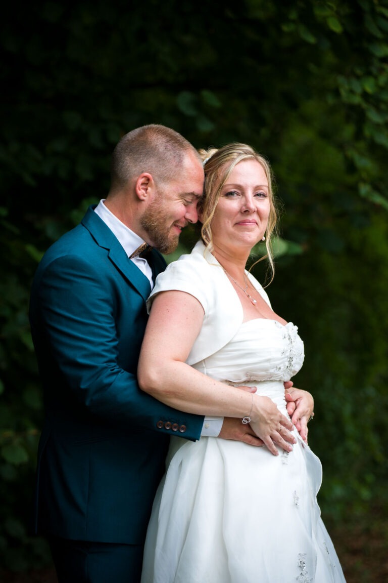 Un couple pose en extérieur dans un cadre verdoyant. Le marié, vêtu d'un costume bleu canard, se tient derrière la mariée, l'enlaçant, les yeux fermés et l'air satisfait. La mariée, vêtue d'une robe de mariée blanche et d'un boléro à manches courtes, sourit doucement tout en regardant la caméra. Ce moment serein capture magnifiquement leur mariage à Bordeaux.