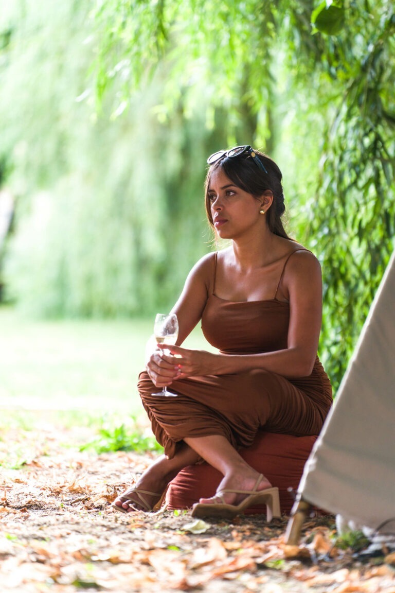 Une femme vêtue d'une robe marron sans manches est assise sur un coussin à l'extérieur, tenant un verre de vin. Elle porte des lunettes de soleil sur la tête et est assise près d'un feuillage vert et d'une structure de tente, avec des feuilles sèches éparpillées sur le sol. L'arrière-plan est flou et semble être un parc ou un jardin luxuriant, peut-être à Bordeaux.