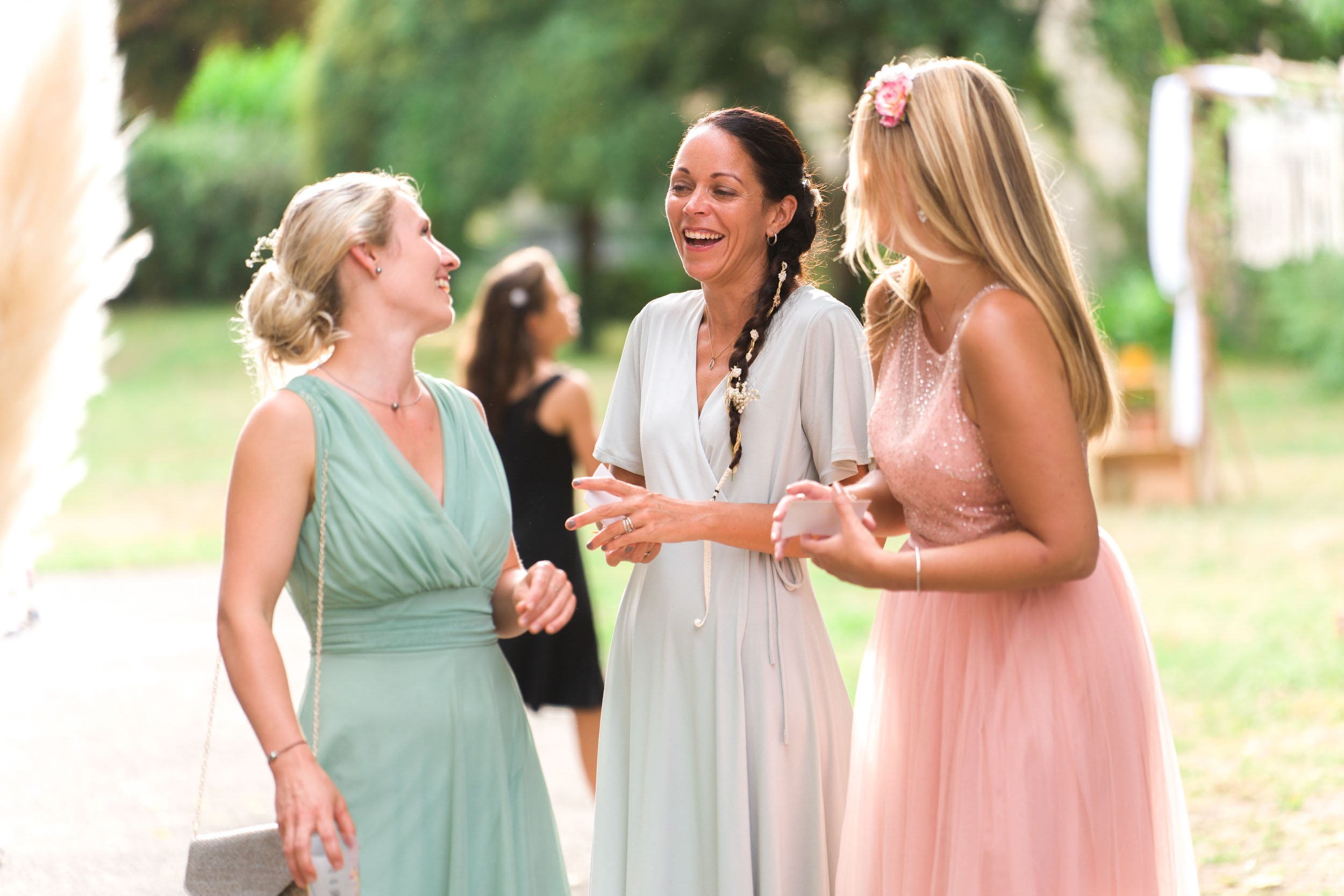 Trois femmes discutent et sourient à l'extérieur, rappelant un moment spontané lors d'un mariage. Elles sont vêtues de robes aux couleurs pastel : l'une en vert, l'autre en gris et l'autre en rose. L'arrière-plan est flou mais suggère un jardin ou un parc. L'atmosphère semble chaleureuse et joyeuse, la lumière naturelle du soleil mettant en valeur la scène.