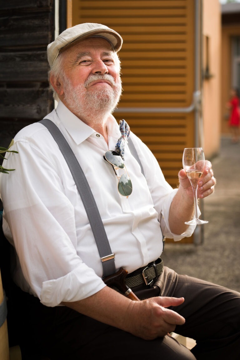 Un homme âgé avec une barbe blanche, portant une chemise blanche, des bretelles grises et une casquette beige, est assis et tient un verre de vin de Bordeaux. Il a des lunettes de soleil accrochées à sa chemise et un foulard autour du cou. Il sourit avec contentement sur un arrière-plan représentant un bâtiment sombre et une porte de garage.