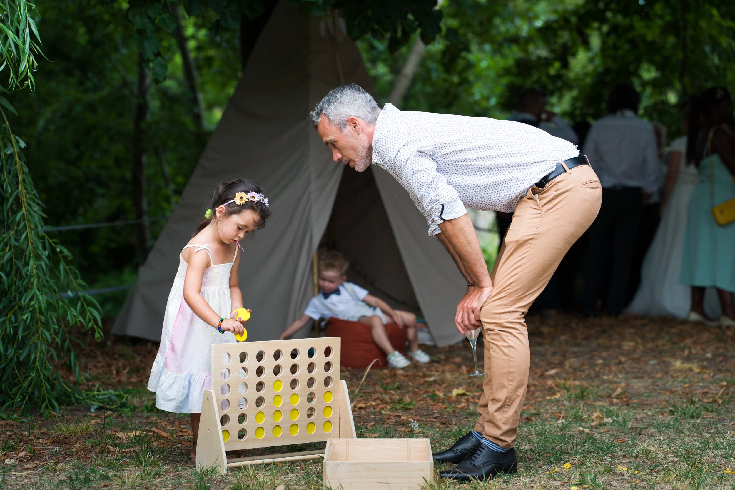 Un homme et une jeune fille jouent à un grand jeu de Puissance 4 en bois en plein air, lors d'un mariage. La jeune fille, vêtue d'une robe blanche et d'un bandeau fleuri, place un disque jaune tandis que l'homme, en chemise blanche et pantalon beige, regarde attentivement. Derrière eux, sous le tipi beige, des capoeiristes se produisent pour les invités en tenue bordeaux.