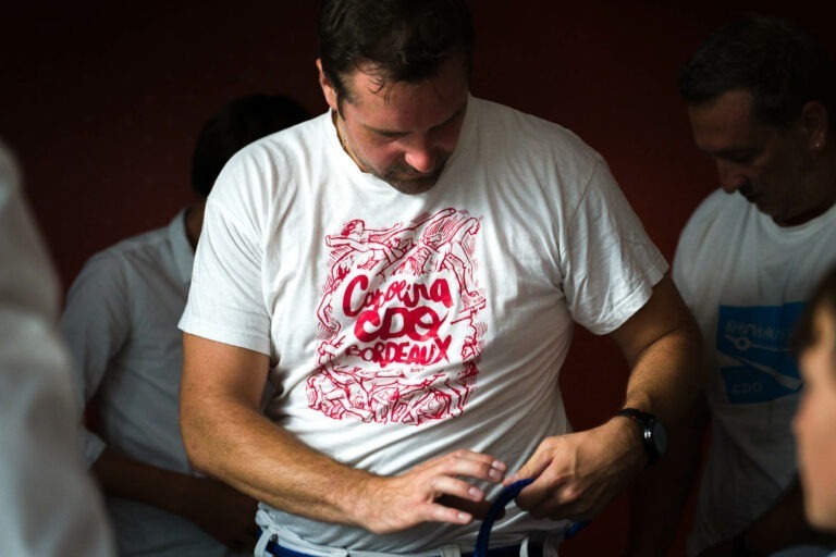 Un homme aux cheveux courts porte un t-shirt blanc avec un imprimé rouge et les mots « Capoeira Bordeaux ». Il ajuste une ceinture bleue autour de sa taille. Une autre personne, vêtue d'une tenue similaire, est légèrement visible dans la pièce faiblement éclairée, se préparant peut-être à célébrer un mariage de capoeiristes.