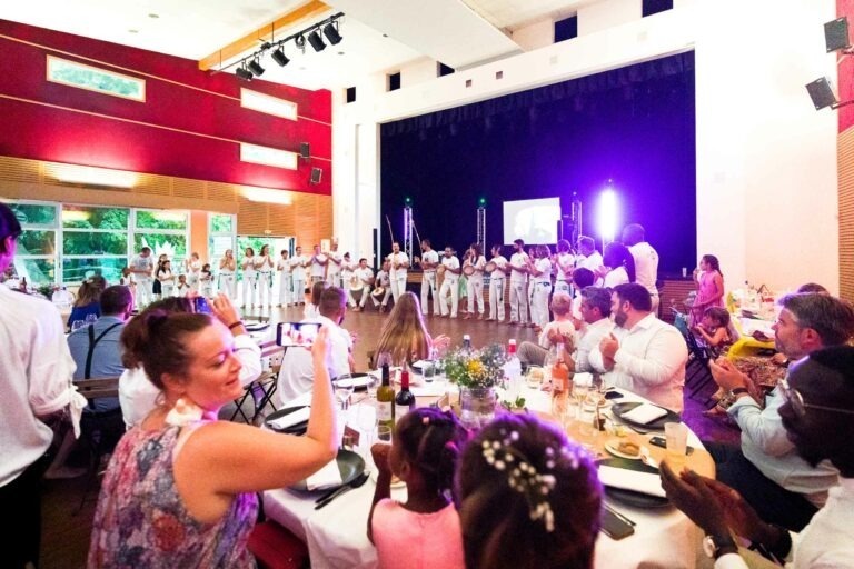 Un événement intérieur animé à Bordeaux capture une performance sur scène avec un groupe de personnes habillées en blanc, probablement des capoeiristes. Le public, assis à des tables rondes ornées de fleurs, regarde attentivement. La salle est bien éclairée avec un décor rouge et blanc. Certains participants prennent des photos et l'ambiance générale est festive.