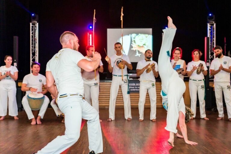 Un groupe de capoeira vêtus d'uniformes blancs se produit sur une scène de Bordeaux. Au premier plan, une personne fait un coup de pied en équilibre sur les mains tout en faisant face à une autre personne dans une position d'arts martiaux. Plusieurs autres personnes jouent d'un instrument et applaudissent, tandis qu'un écran à l'arrière-plan diffuse une image, créant un mariage captivant de mouvement et de rythme.