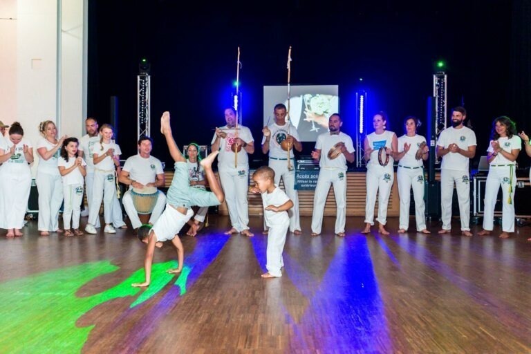 Un groupe de personnes vêtues d'uniformes blancs se tient en demi-cercle sur un plancher en bois, jouant d'un instrument de musique et applaudissant. Deux enfants exécutent une capoeira au centre, l'un d'eux faisant le poirier tandis que l'autre se tient en équilibre sur une main. Les capoeiristes apportent de la vivacité à ce qui ressemble à une célébration de mariage, illuminée par les lumières de la scène.