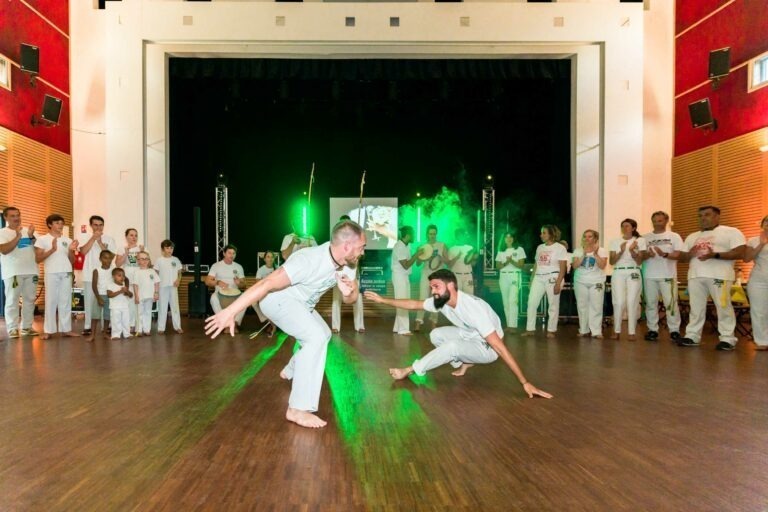 Deux individus vêtus de blanc exécutent une capoeira sur un plancher en bois, entourés d'un cercle de spectateurs, certains applaudissant et d'autres jouant d'un instrument. L'arrière-plan présente une scène avec des lumières vertes et de la fumée. L'atmosphère énergique rappelle la forme d'art culturel vibrante que l'on retrouve lors des mariages capoeiristes à Bordeaux.