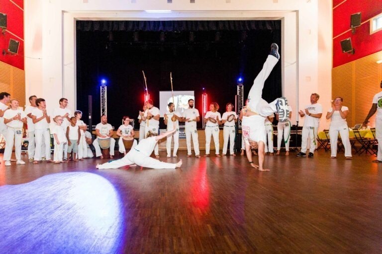 Un groupe de personnes, vêtues de la tenue blanche traditionnelle de capoeira, forment un cercle dans un gymnase éclairé lors d'un mariage. Deux capoeiristes exécutent des mouvements fascinants au centre ; l'un fait un salto tandis que l'autre exécute un mouvement bas et ample. Les spectateurs applaudissent et acclament.