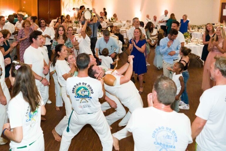 Un groupe de capoeiristes dynamiques se livre à une performance de capoeira lors d'une soirée à Bordeaux. Les artistes en tenue blanche, certains marqués du sigle « Cordão de Ouro Capoeira », interagissent avec énergie tandis qu'une foule de spectateurs, dont des enfants et des adultes, les encouragent. Une personne joue du tambourin en arrière-plan.