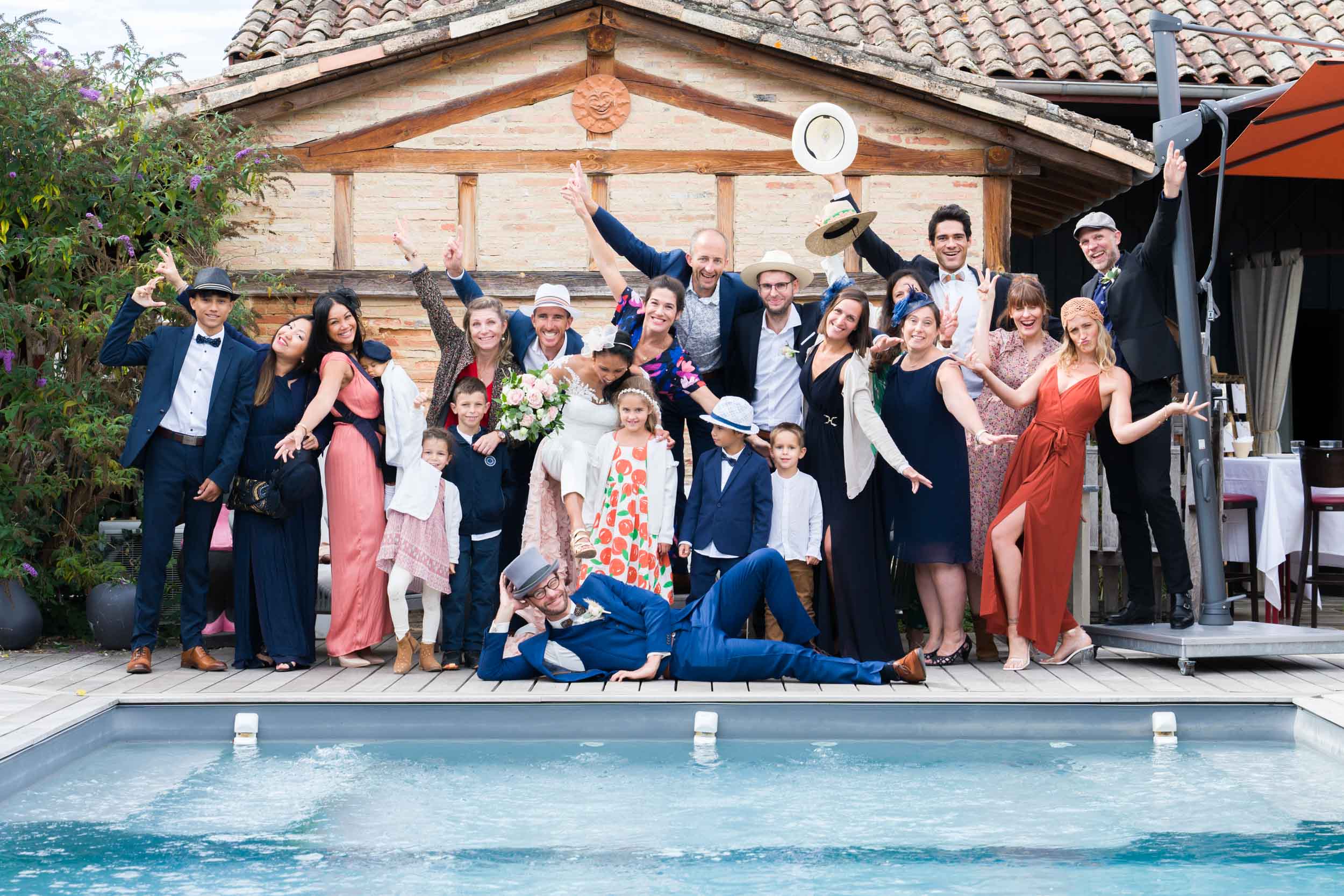 Une photo de groupe joyeuse au bord de la piscine capture des personnes bien habillées de différents âges posant de manière ludique, certaines levant les bras ou faisant des gestes. Sur une terrasse en bois devant un bâtiment rustique en bois, la scène dégage un air festif rappelant un mariage simple et élégant, avec des vêtements vibrants et des expressions joyeuses évidentes parmi tous les participants.