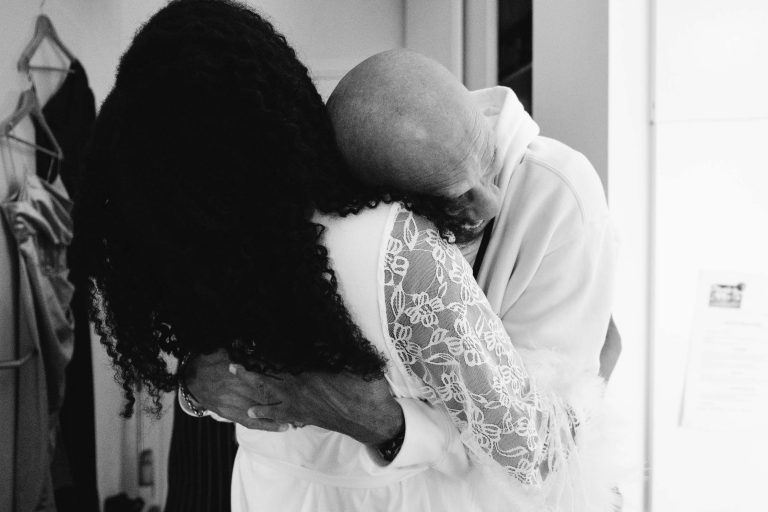 Une photo en noir et blanc de deux personnes qui s'embrassent. La personne de gauche a les cheveux longs et bouclés et porte un chemisier à manches en dentelle. La personne de droite, chauve et portant un sweat à capuche de couleur claire, pose sa tête sur l'épaule de l'autre. Des vêtements de Mariage Audenge pendent à l'arrière-plan.