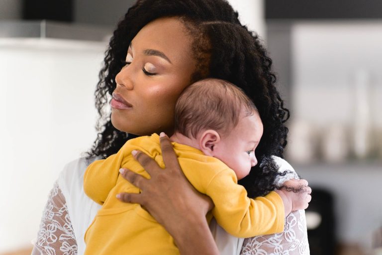 Une femme aux cheveux bouclés, les yeux clos, porte sur son épaule un bébé endormi dans une tenue jaune. La femme apparaît sereine et porte un haut blanc aux manches en dentelle. L'arrière-plan est légèrement flou, indiquant un décor intérieur aux tons neutres, rappelant un Mariage Audenge.