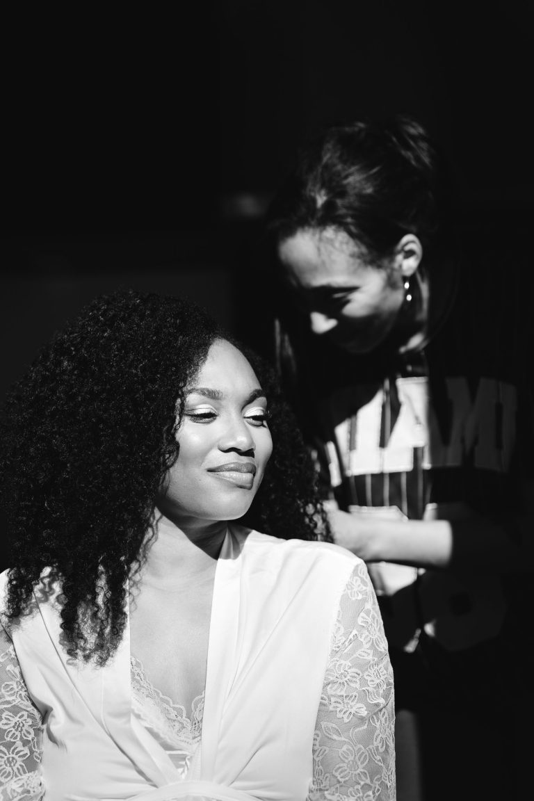 Une photo en noir et blanc montre une femme aux cheveux bouclés, vêtue d'un vêtement clair, souriant tout en regardant sur le côté. Une autre personne aux cheveux bouclés se penche en arrière-plan, engagée dans une activité concentrée, partiellement dans l'ombre, portant une chemise sombre avec des lettres visibles. Ce moment capture l'essence candide de Mariage Audenge.