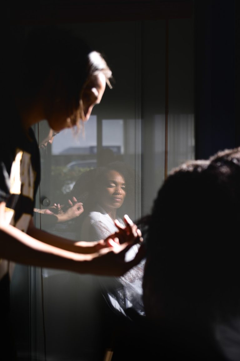 Dans une pièce faiblement éclairée, une femme coiffe les cheveux d'une autre personne pour un mariage Audenge à venir. La femme qui se fait coiffer est assise, regardant son reflet dans la fenêtre, qui capture son visage souriant. La lumière du soleil met en valeur à la fois son visage et la main du coiffeur qui lui peigne doucement les cheveux.