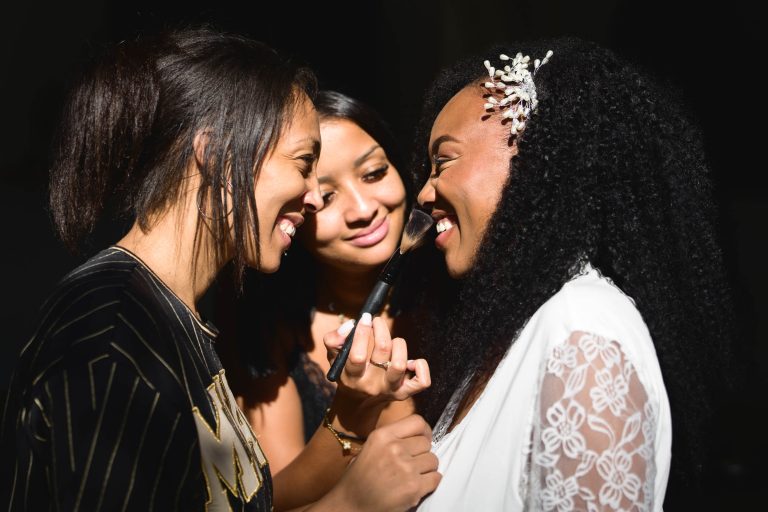 Trois femmes sourient et interagissent étroitement. Une femme à gauche se maquille avec un pinceau sur la joue de la femme à droite, qui a les cheveux bouclés ornés d'épingles décoratives. La femme au milieu les observe en souriant, se préparant peut-être à une célébration du Mariage Audenge. L'arrière-plan est sombre, ce qui fait ressortir les femmes.