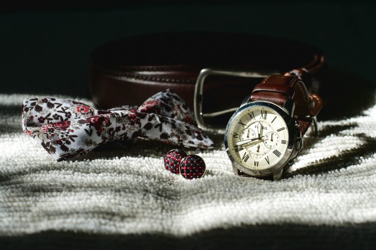 Une montre-bracelet avec bracelet en cuir marron, un nœud papillon à motifs floraux, une paire de boutons de manchette rouges et une ceinture en cuir marron avec boucle argentée de la collection Mariage Audenge sont disposés sur une surface texturée et claire. La composition est illuminée par une lumière douce et directionnelle, créant une atmosphère sophistiquée.