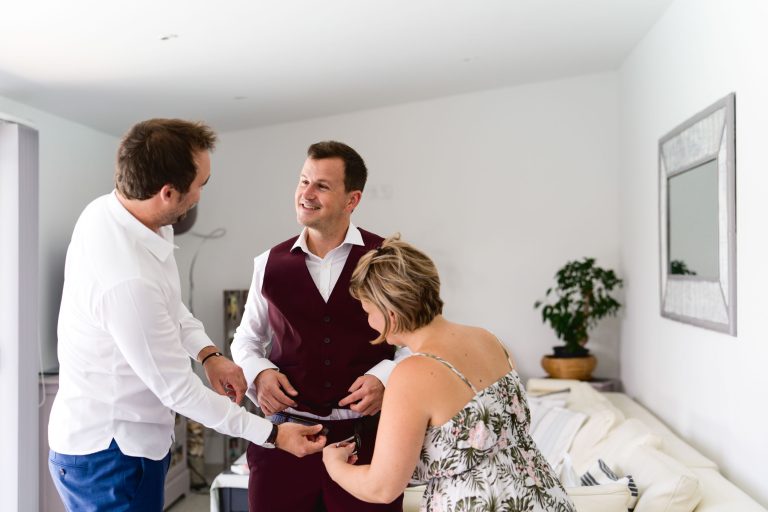 Un homme portant un gilet bordeaux et une chemise blanche, se préparant probablement pour le mariage Audenge, se tient dans une pièce lumineuse. Il est assisté d'une femme portant une robe fleurie et d'un homme portant une chemise blanche et un pantalon bleu. Ils ajustent sa tenue. La pièce a des murs blancs, un canapé, une plante en pot et un grand miroir.