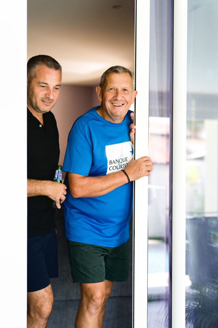 Deux hommes sourient en franchissant une porte coulissante en verre partiellement ouverte. L'homme devant, vêtu d'un t-shirt bleu et d'un short foncé, s'agrippe au cadre de la porte. L'homme derrière, vêtu d'une chemise noire et d'un short foncé, tient une bouteille verte. Ils semblent détendus et joyeux dans leur décor après avoir apprécié le Mariage Audenge.