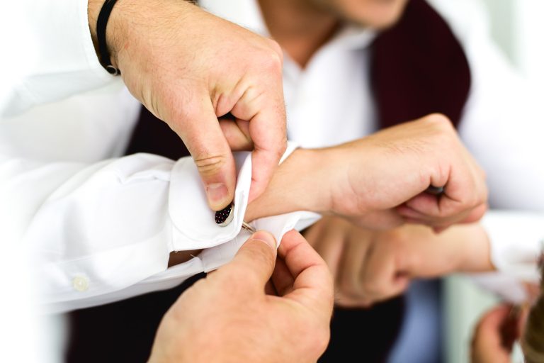 Gros plan d'une personne attachant des boutons de manchette sur une manche de chemise blanche. Les mains d'une autre personne aident à ajuster la manchette. Les deux individus portent des chemises blanches ; l'un d'eux est également vu avec un gilet foncé. L'image, probablement tirée d'un mariage Audenge, se concentre sur les mains et leur tâche, soulignant la préparation d'un événement officiel.