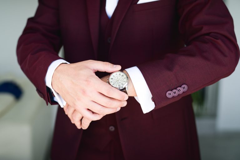 Une personne vêtue d'un costume bordeaux avec une chemise blanche ajuste une montre-bracelet en argent. La veste du costume a quatre boutons assortis sur le poignet. La scène se déroule à l'intérieur avec un arrière-plan flou, reflétant l'élégance et le style qui conviennent aux tenues et accessoires Mariage Audenge.