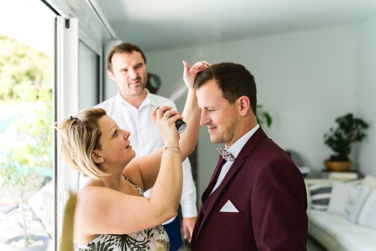 Une femme vaporise du produit capillaire sur les cheveux d'un homme, vêtu d'un costume marron et d'un nœud papillon, se préparant probablement pour un mariage Audenge. Un autre homme en chemise blanche se tient à proximité et observe. Ils sont à l'intérieur, avec de la lumière naturelle entrant par une grande fenêtre et des plantes visibles en arrière-plan.