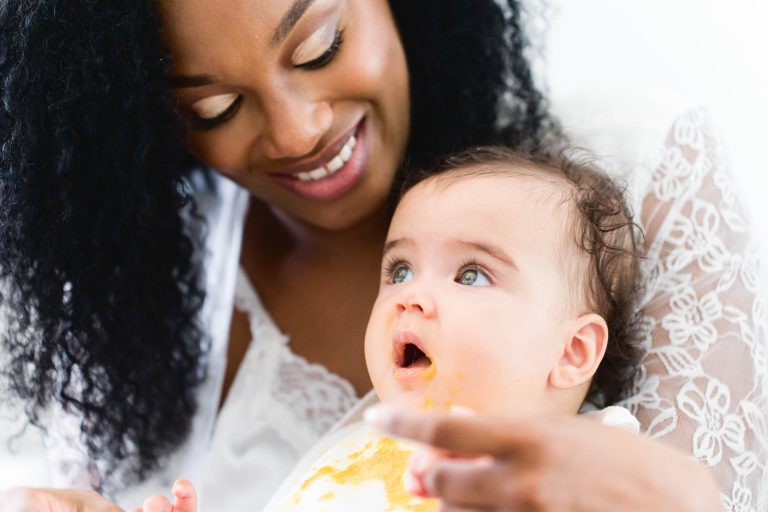 Une femme aux cheveux bouclés sourit en tenant dans ses bras un bébé aux cheveux châtain clair. Le bébé, la bouche étalée de nourriture, regarde vers le haut avec curiosité. La femme, vêtue d'une tenue en dentelle blanche rappelant un mariage de Mariage Audenge, les baigne tous les deux dans une douce lumière naturelle.