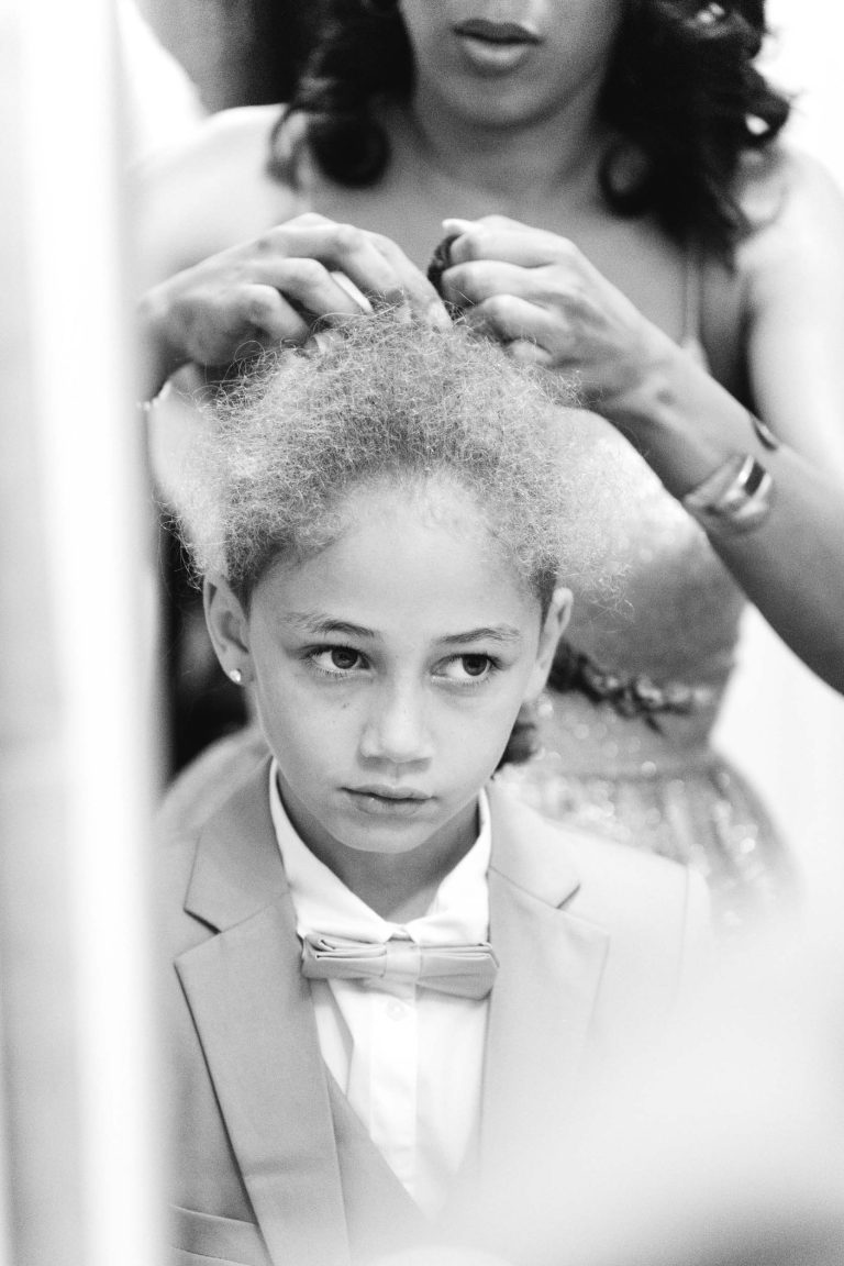 Photographie en noir et blanc d'un jeune enfant portant un costume et un nœud papillon, qui se fait coiffer par une femme adulte. L'enfant a une expression pensive tandis que la femme, partiellement visible, ajuste les cheveux de l'enfant en prévision de ce qui ressemble à un événement de mariage Audenge.